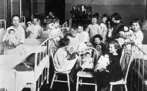 A group of children holding dolls in a room with beds, possibly a hospital or orphanage setting.