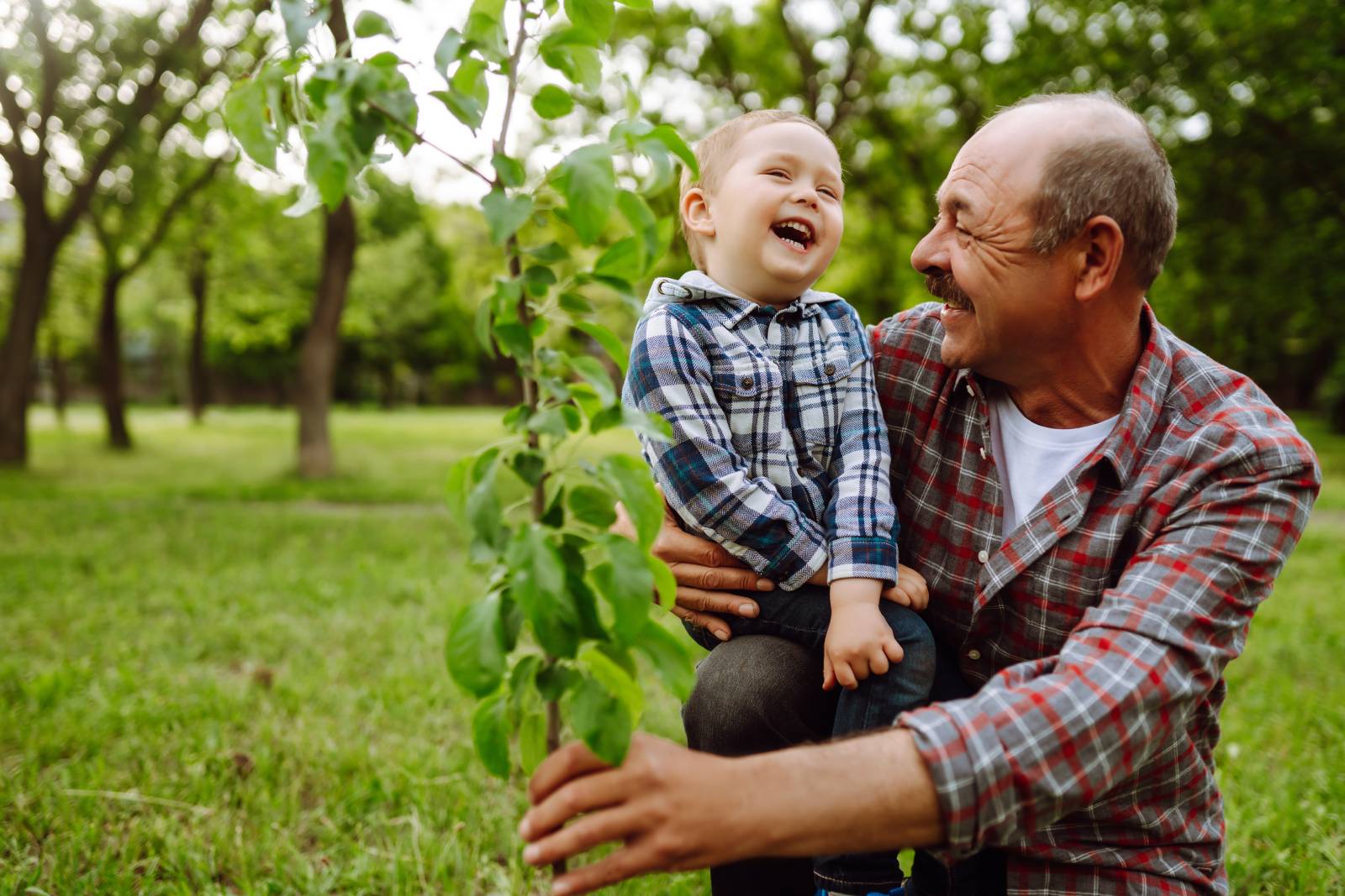 An elderly man and a young child share a joyful moment outdoors in a lush, green park setting.