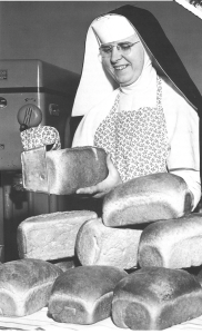A nun is holding a loaf of bread, surrounded by other loaves on a table.