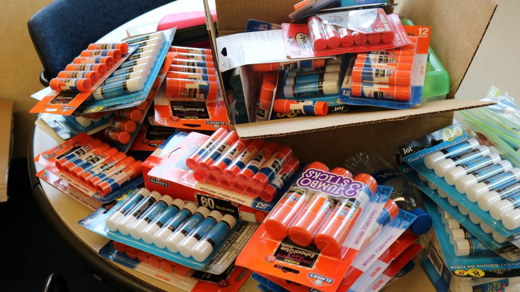 A large assortment of glue sticks and school supplies is displayed, related to a 2026 back to school drive.