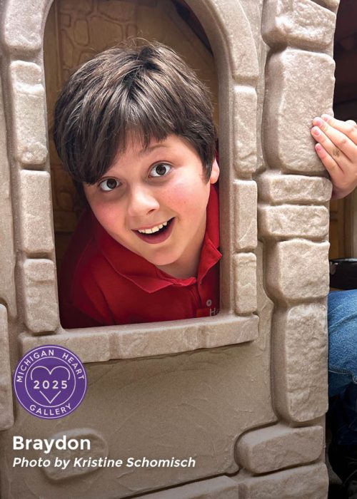 A child smiles through a playhouse window. Label shows "Michigan Heart Gallery 2025" by Kristine Schomisch.