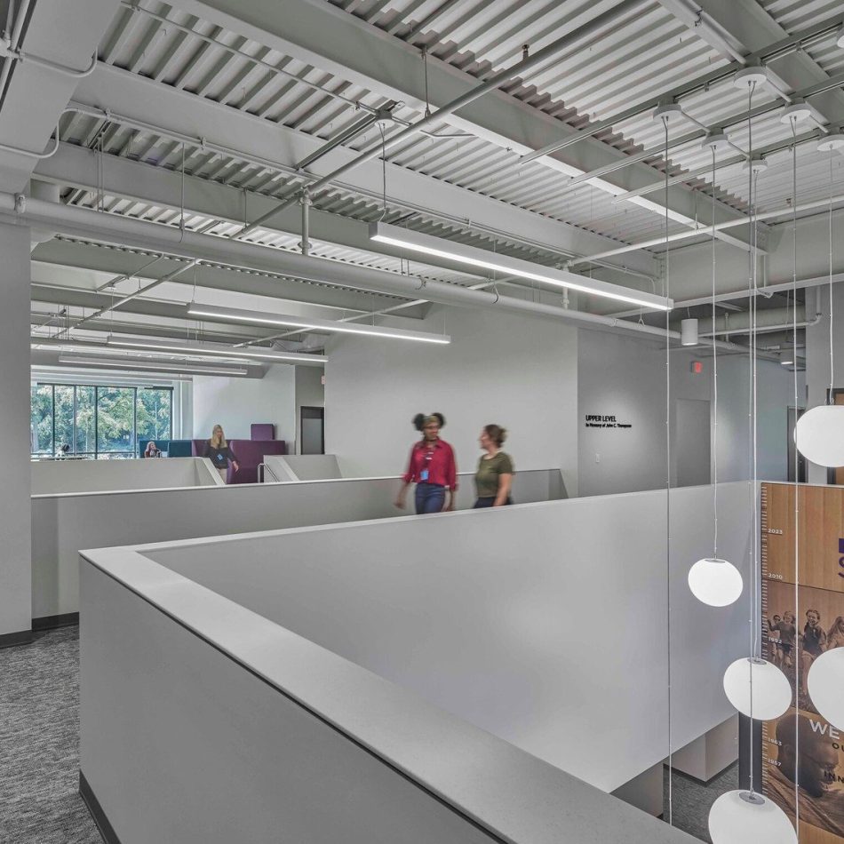 A modern office interior with people walking, featuring open workspaces and a visible ceiling structure.
